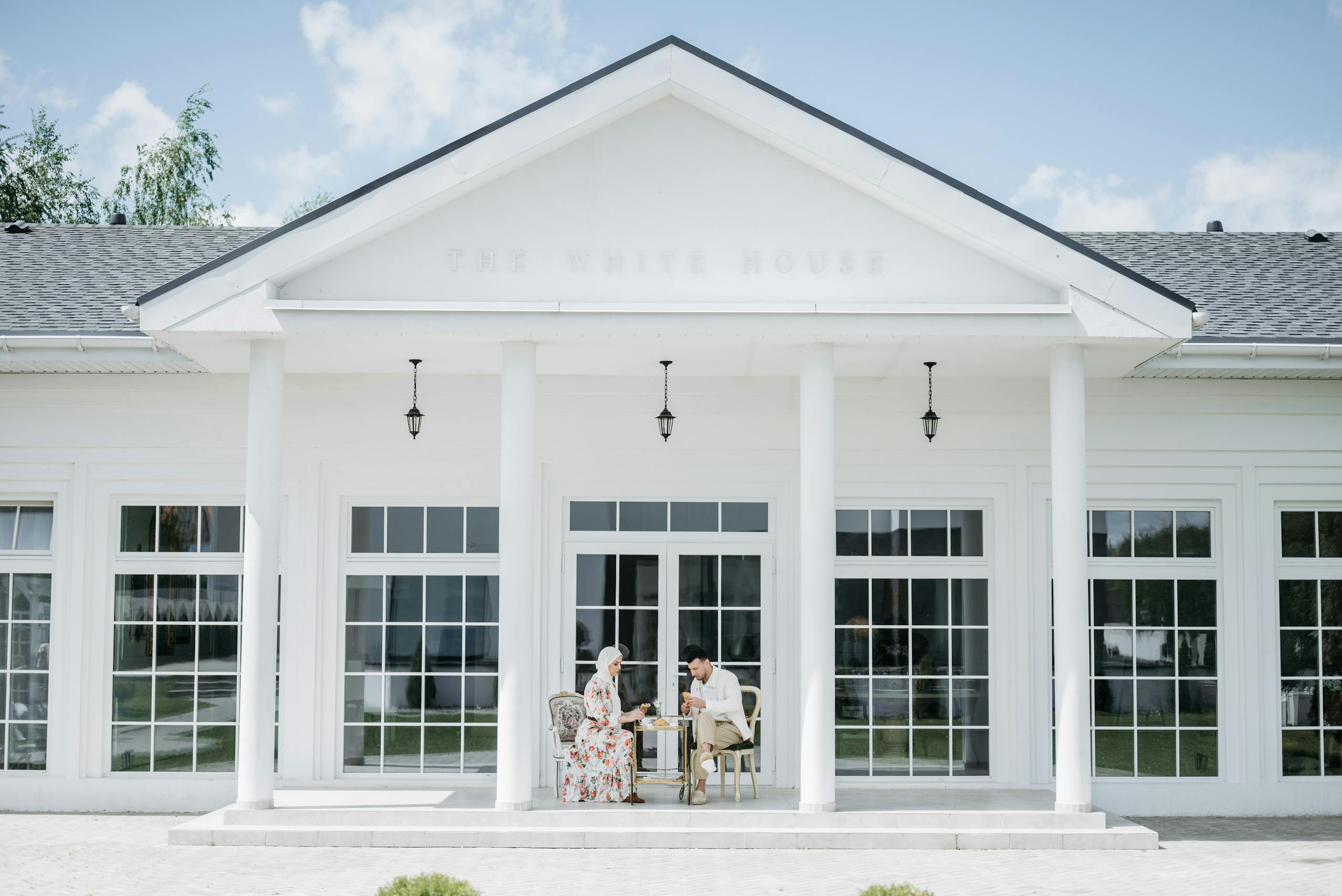 A couple enjoying a sunny day in front of a luxurious white mansion.