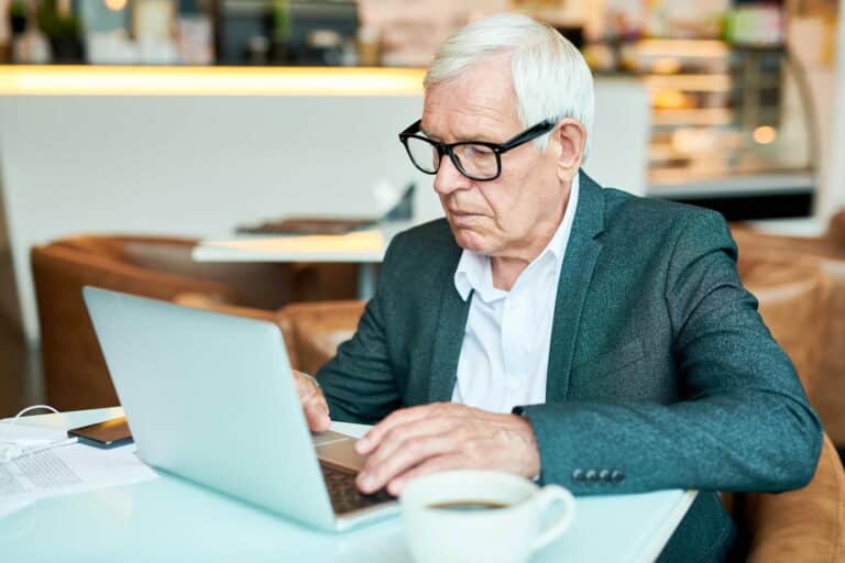 An older man wearing glasses is focused on using a laptop at a desk.
