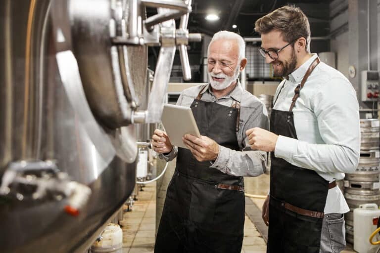 Two men in aprons stand in front of a large tank, preparing for a task related to food or beverage production.