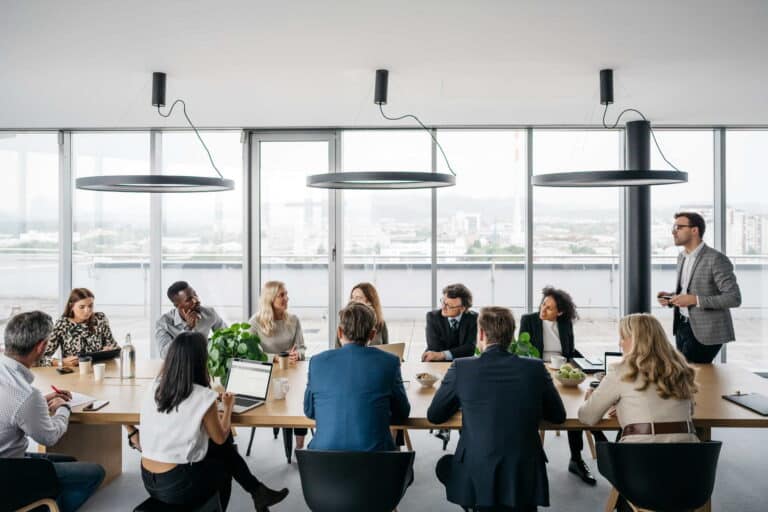 A group of business professionals engaged in discussion around a conference table in a modern meeting room.