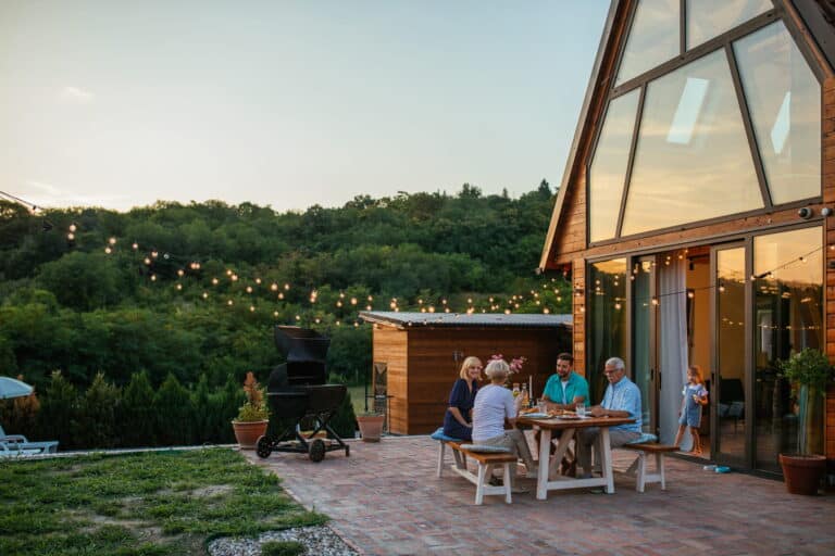 A family enjoys a meal outside at a table, with a grill in the background, surrounded by greenery