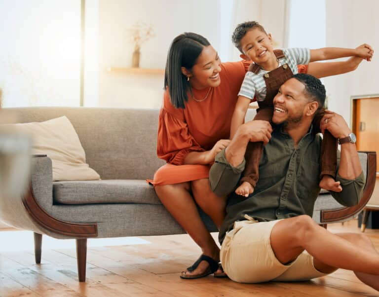 A family of four sitting together on the floor in a cozy living room, surrounded by toys and cushions.