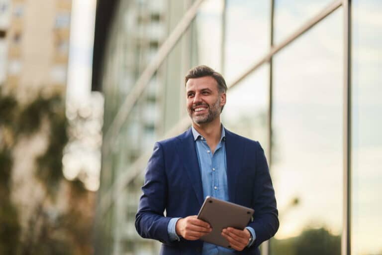 Smiling businessman holding a tablet, standing in a bustling city environment.
