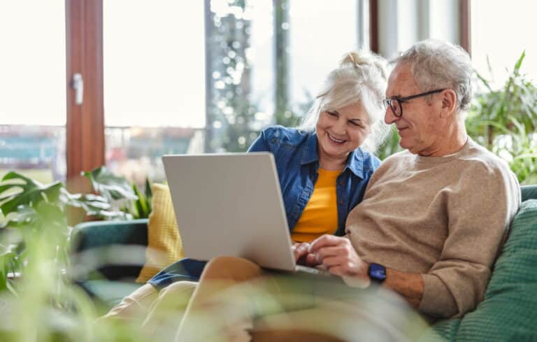 An older couple sits together on a couch, using a laptop and sharing a moment of connection and engagement.