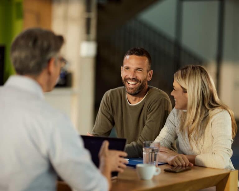 A man and woman sit at a table, focused on a laptop, engaged in discussion or collaboration.