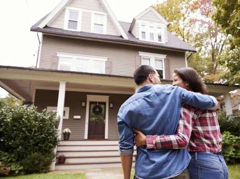 A couple stands together in front of a house, smiling and enjoying their time outdoors.